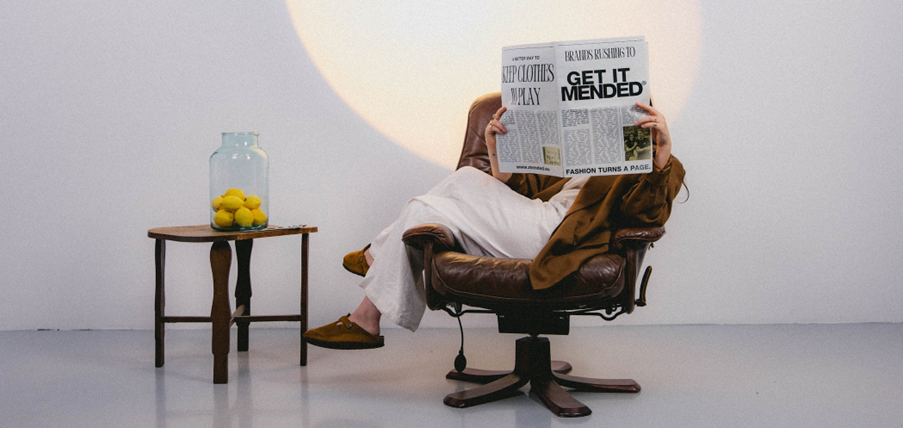 Een persoon die achterover in een stoel zit en een krant vasthoudt voor het gezicht. Op de krant staan titels als 'get it mended', wat refereert aan kledingreparatie.